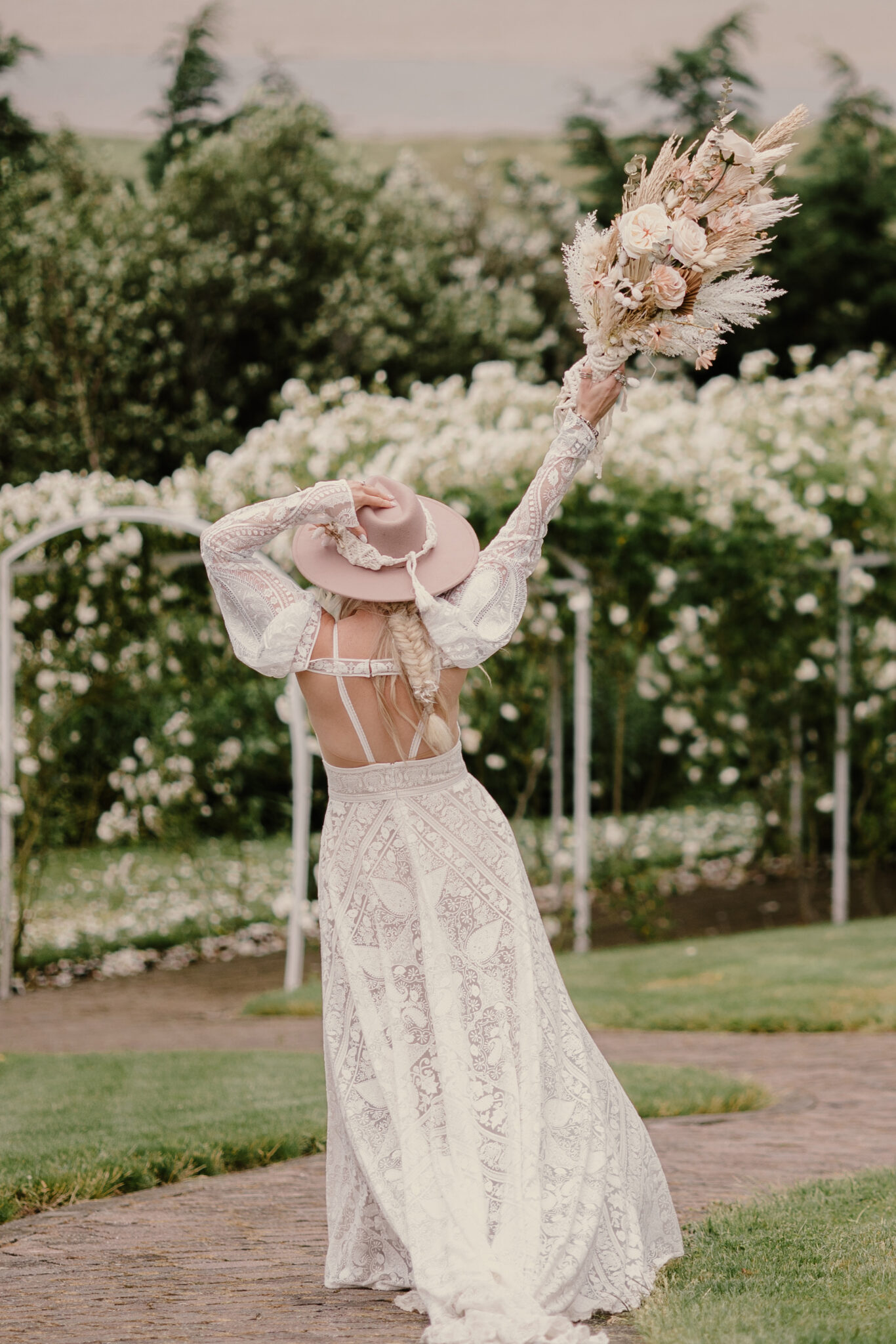 Bride in a vintage lace wedding dress and hat raises her bouquet with blooming rose arch in the background