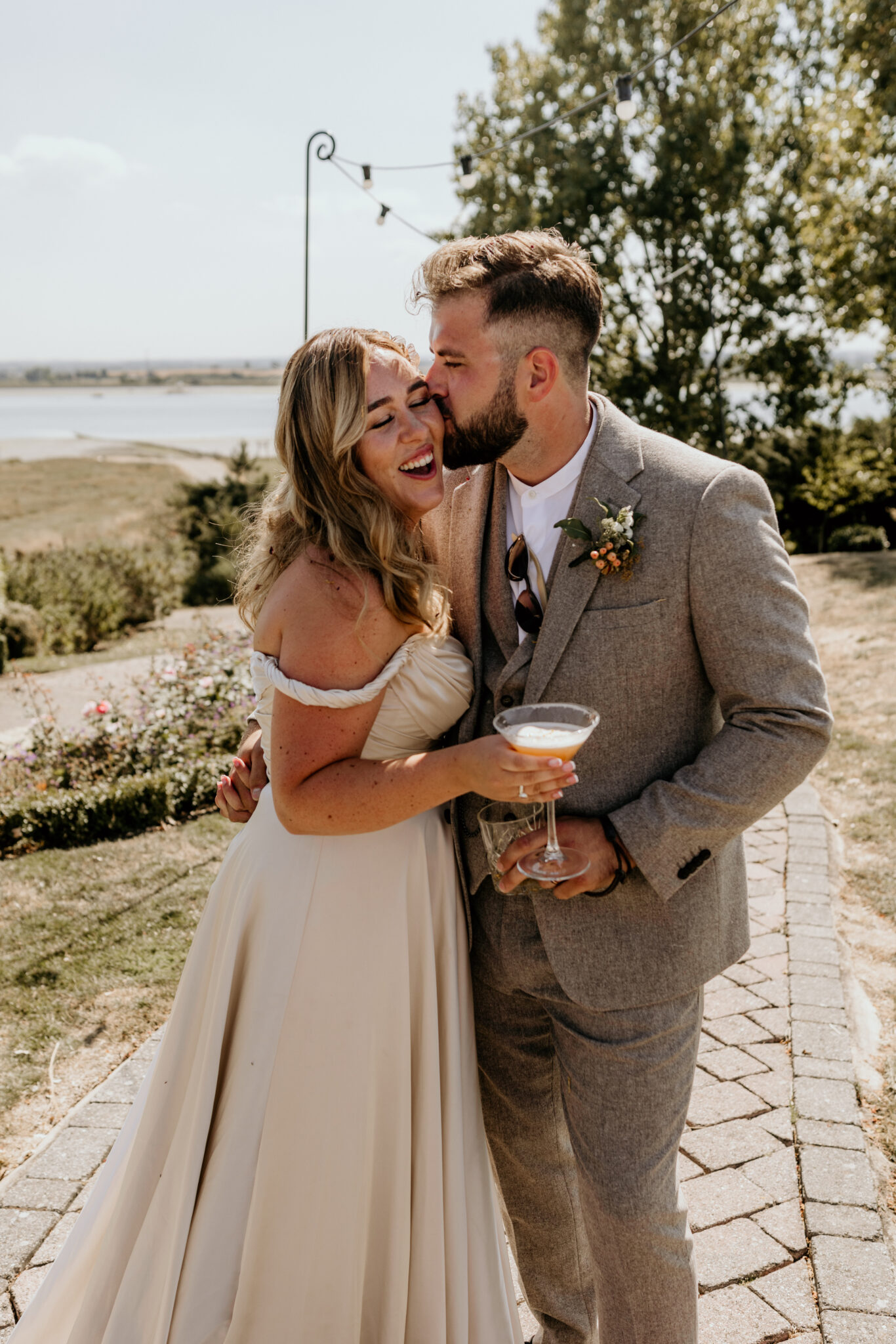 Bride holding a cocktail gets a kiss from her husband in a Kent wedding garden