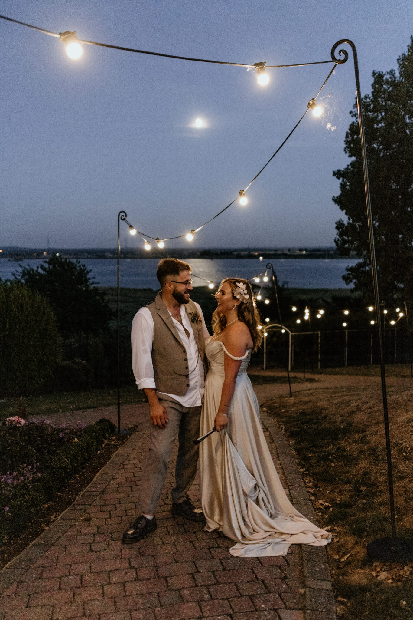 Wedding couple in a waterside wedding garden at twilight beneath glowing festoon lights