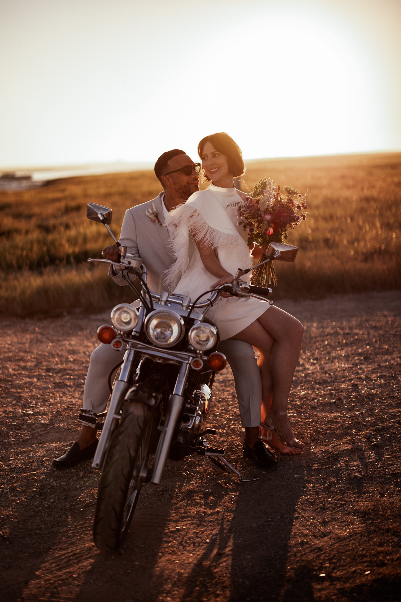Glamorous couple sit on a motorbike at twilight at a waterside wedding venue