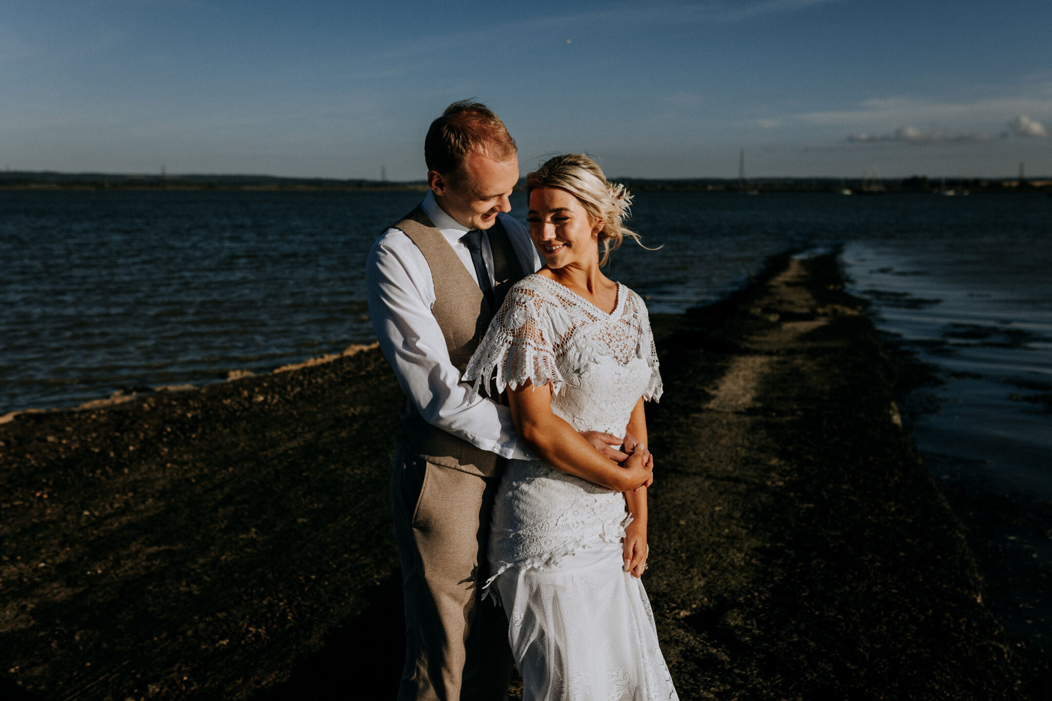 Bride in lace dress held by her husband beside the Swale estuary in Kent