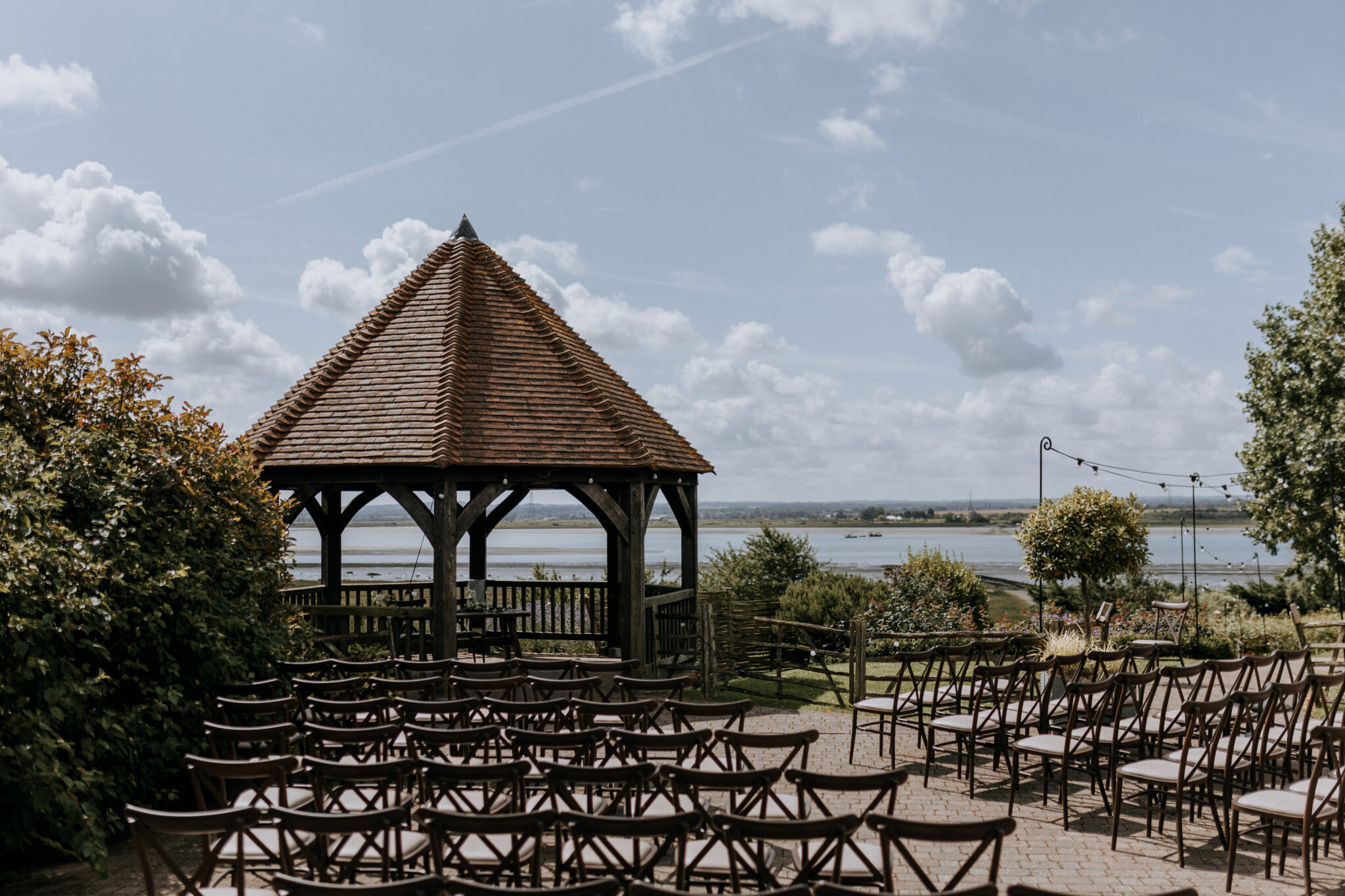 Outdoor wedding ceremony setup on a sunlit terrace with oak gazebo overlooking the water