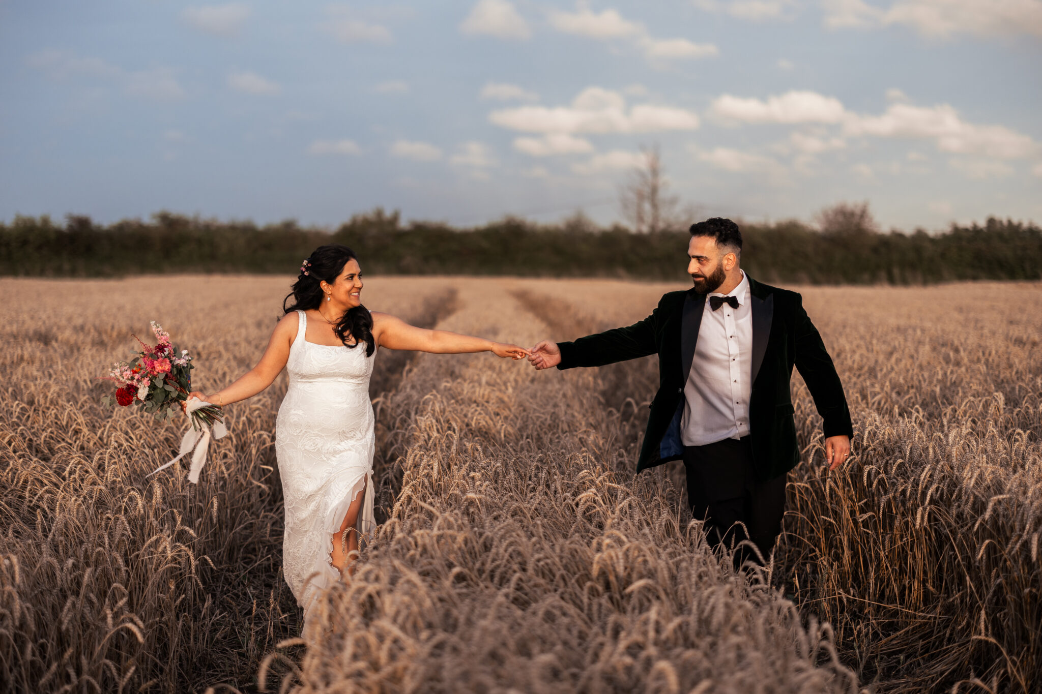Bride and groom hold hands in a golden wheat field at a rustic barn wedding venue in Kent