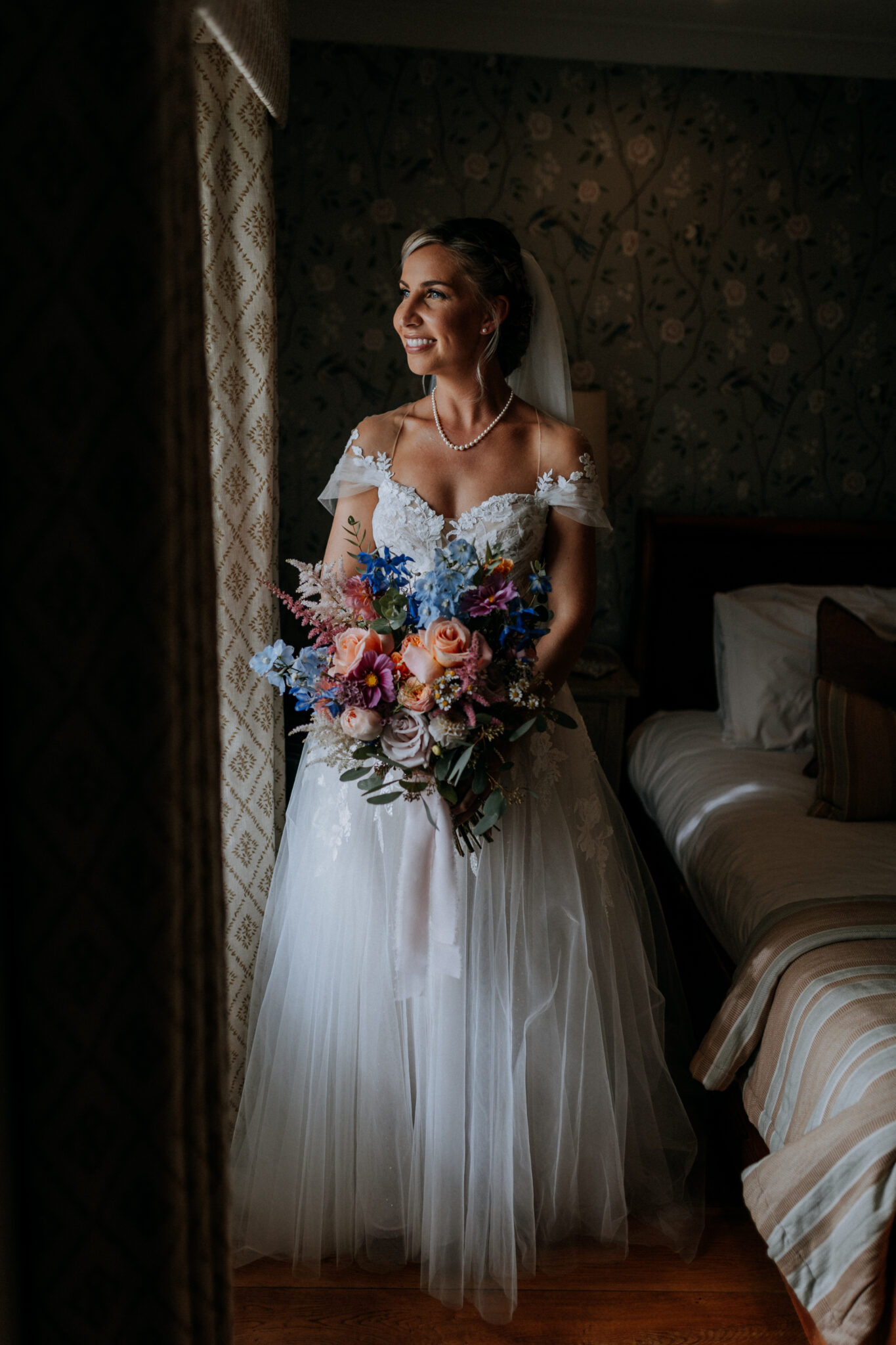 Beaming bride with large and colourful bouquet stands by the window of her wedding venue accommodation