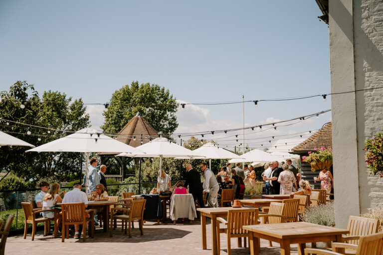 Weddings guests gather under parasols and festoon lights at sunny wedding venue drinks reception