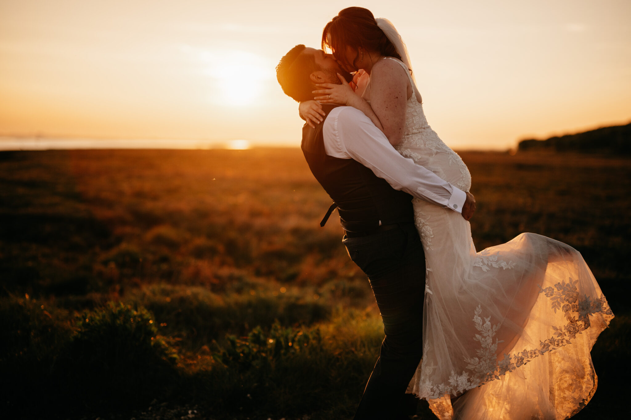 Romantic twilight kiss as groom lifts bride among wetlands of countryside wedding venue