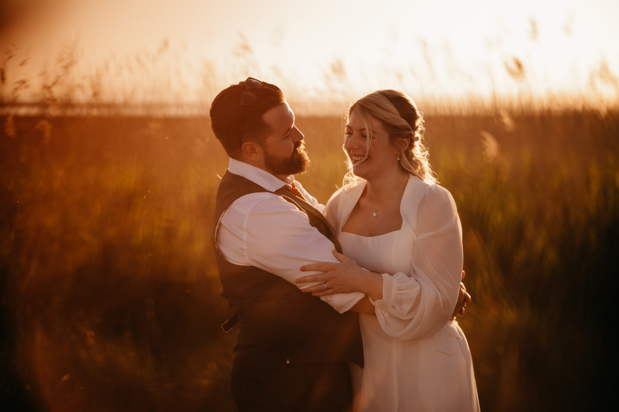 Wedding couple laughs and embraces, backed by swaying grass at twilight