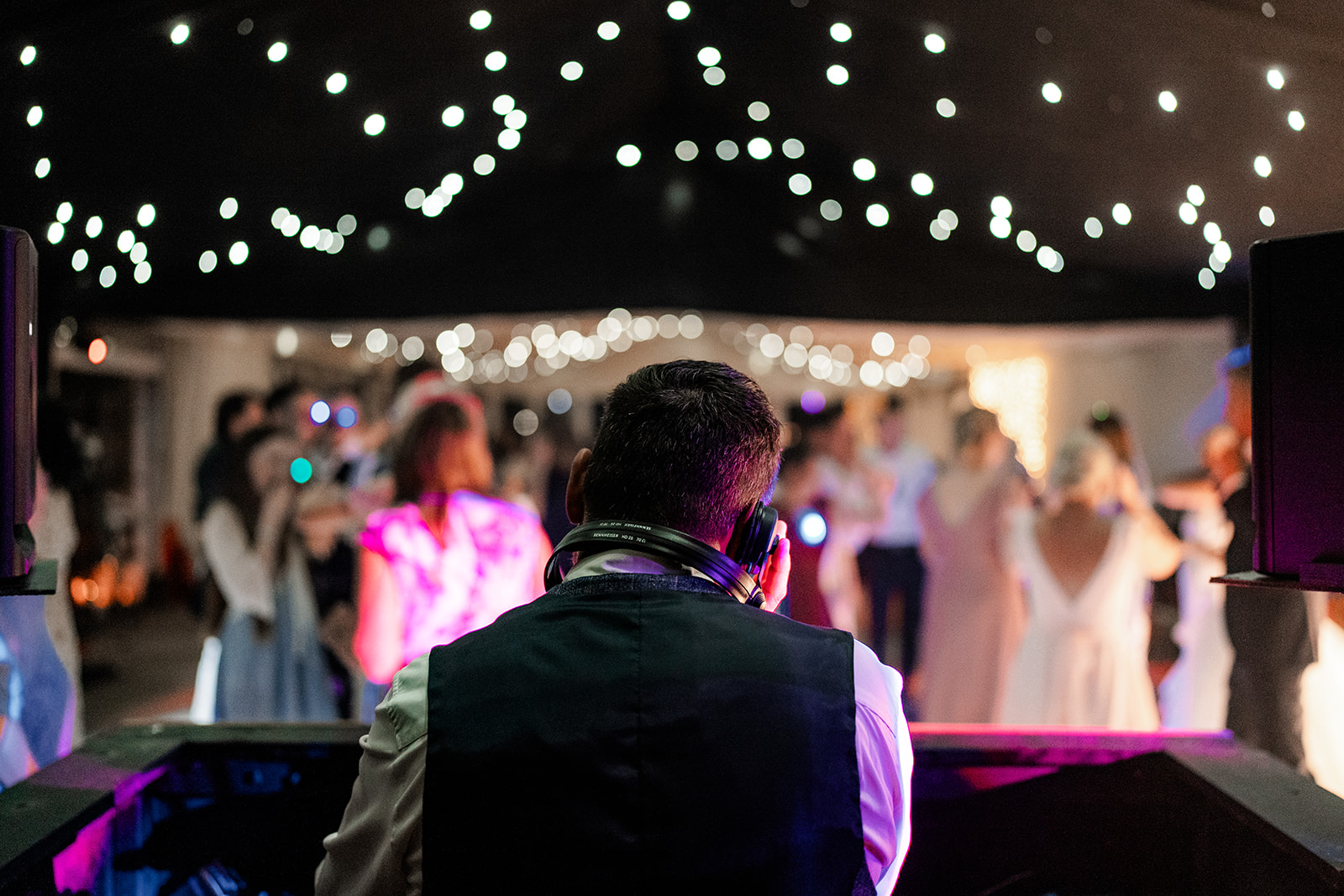 From behind, a Wedding DJ operating the decks to a packed dancefloor, beneath fairylights
