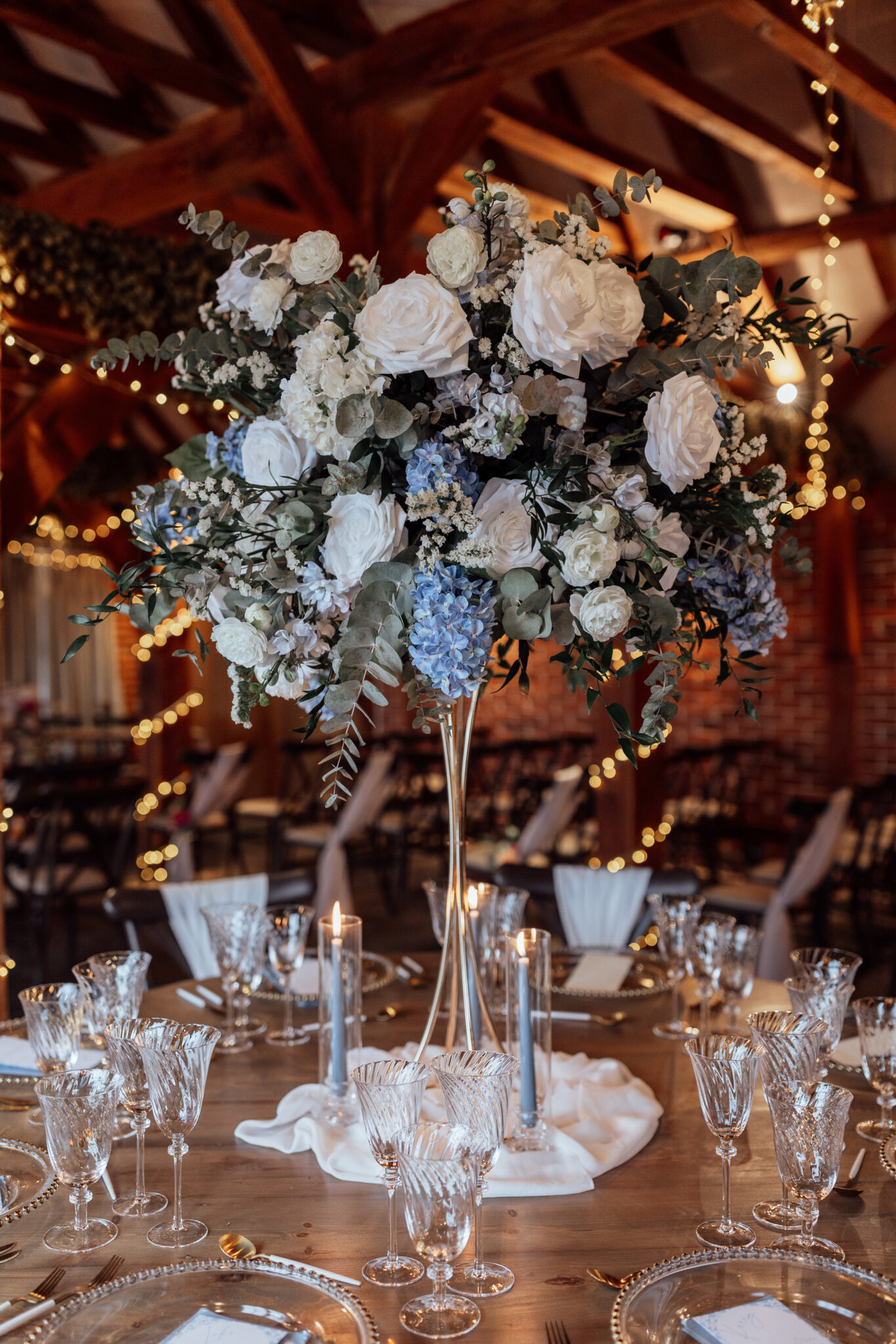Wedding Breakfast table in a barn, with tall glass vase of white and blue flowers