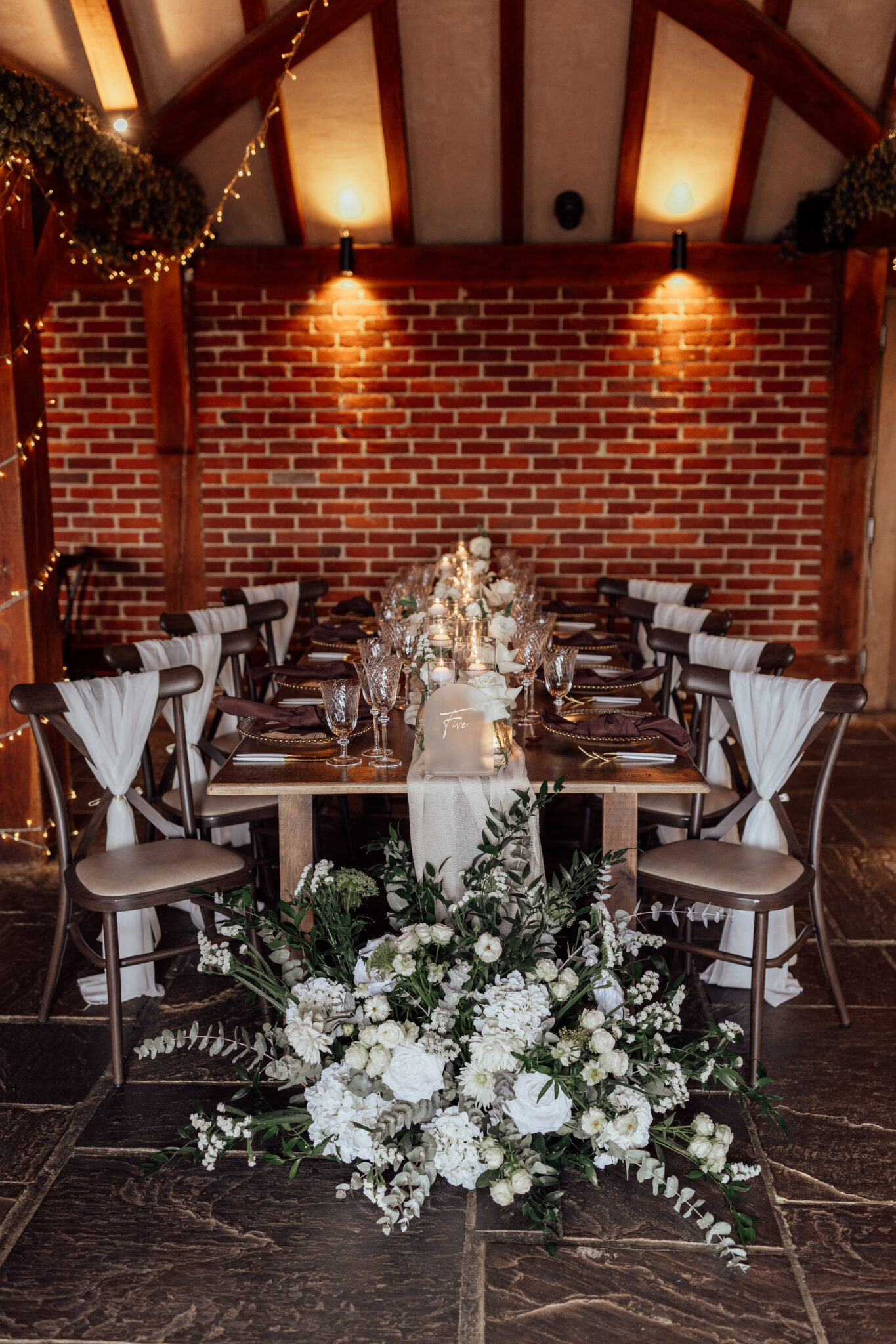 Rectangular wedding breakfast table styled in a rustic barn with white and green florals, table runner and char sashes
