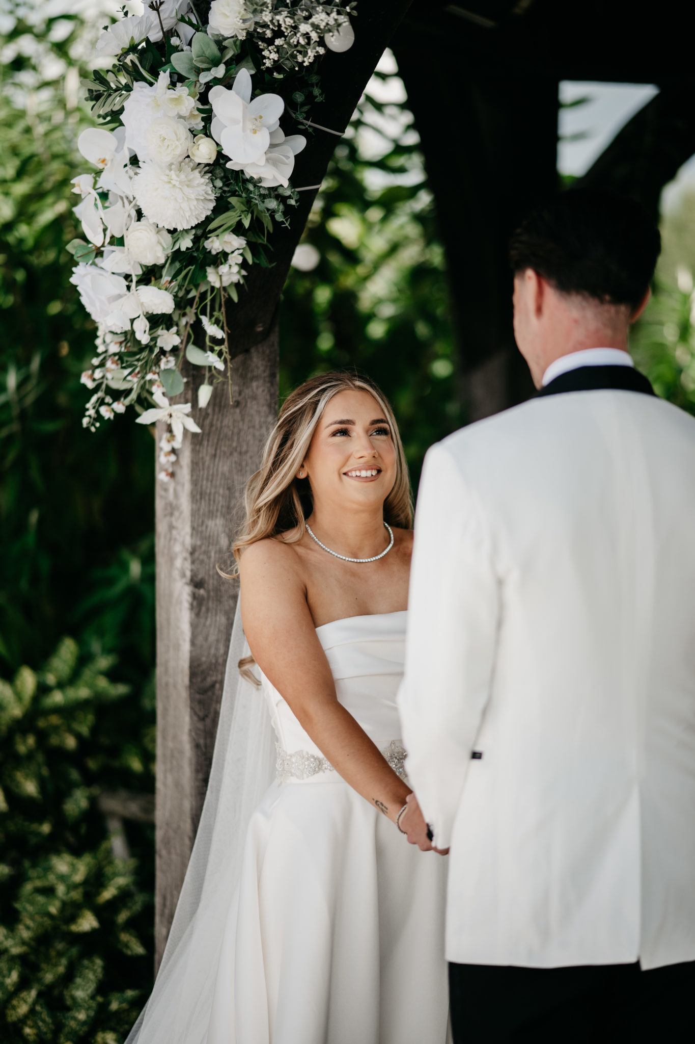 Bride and groom join hands during their outdoor wedding ceremony, beneath a gazebo dressed in white flowers