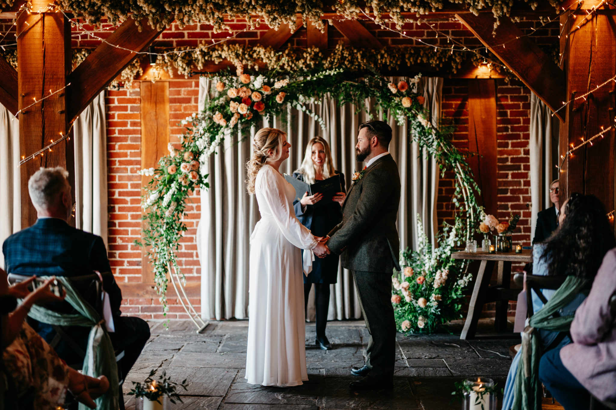 Bride and groom hold hands at the end of a barn wedding aisle, in front of a floral moongate