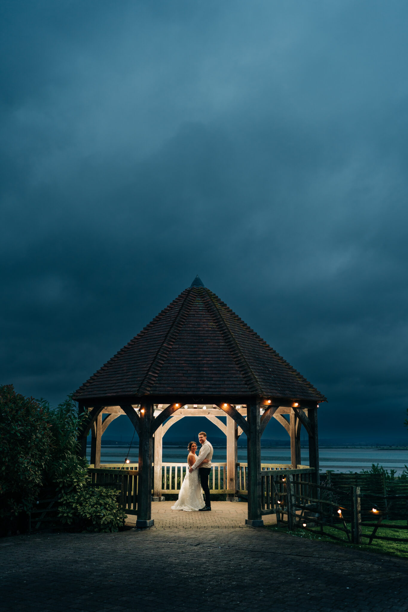 Newlyweds illuminated beneath a waterside gazebo at nightfall